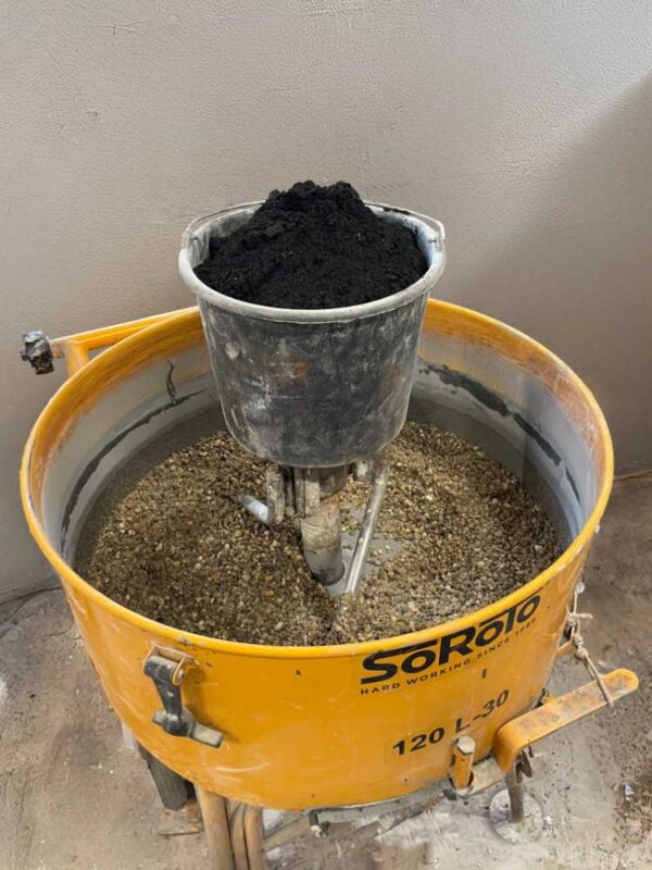 Das Bild zeigt einen geöffneten Betonmischer, gefüllt mit einer Gesteinskörnung als Bestandteil der Betonmischung. Im Zentrum steht ein Metalleimer, randvoll mit feiner, schwarzer Biokohle – einem zukunftsweisenden Zusatzstoff für klimafreundlichere Betone. Mit dem Einsatz von Biokohle in der Betonproduktion beschreitet Betoniu im Rahmen eines durch das "Unternehmen Revier" geförderten Projekts innovative Wege, den CO₂-Fußabdruck seiner Betonmöbel weiter zu reduzieren. Biokohle "versenkt" CO₂ im Zement – ein Beitrag zum Klimaschutz.