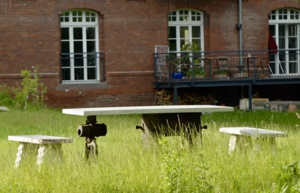 Auf einer großzügigen Grünfläche steht ein Tisch mit Betoniu-Tischplatte. DIese liegt auf industriellen Artefakten auf und wird von Betonbänken ergänzt.
